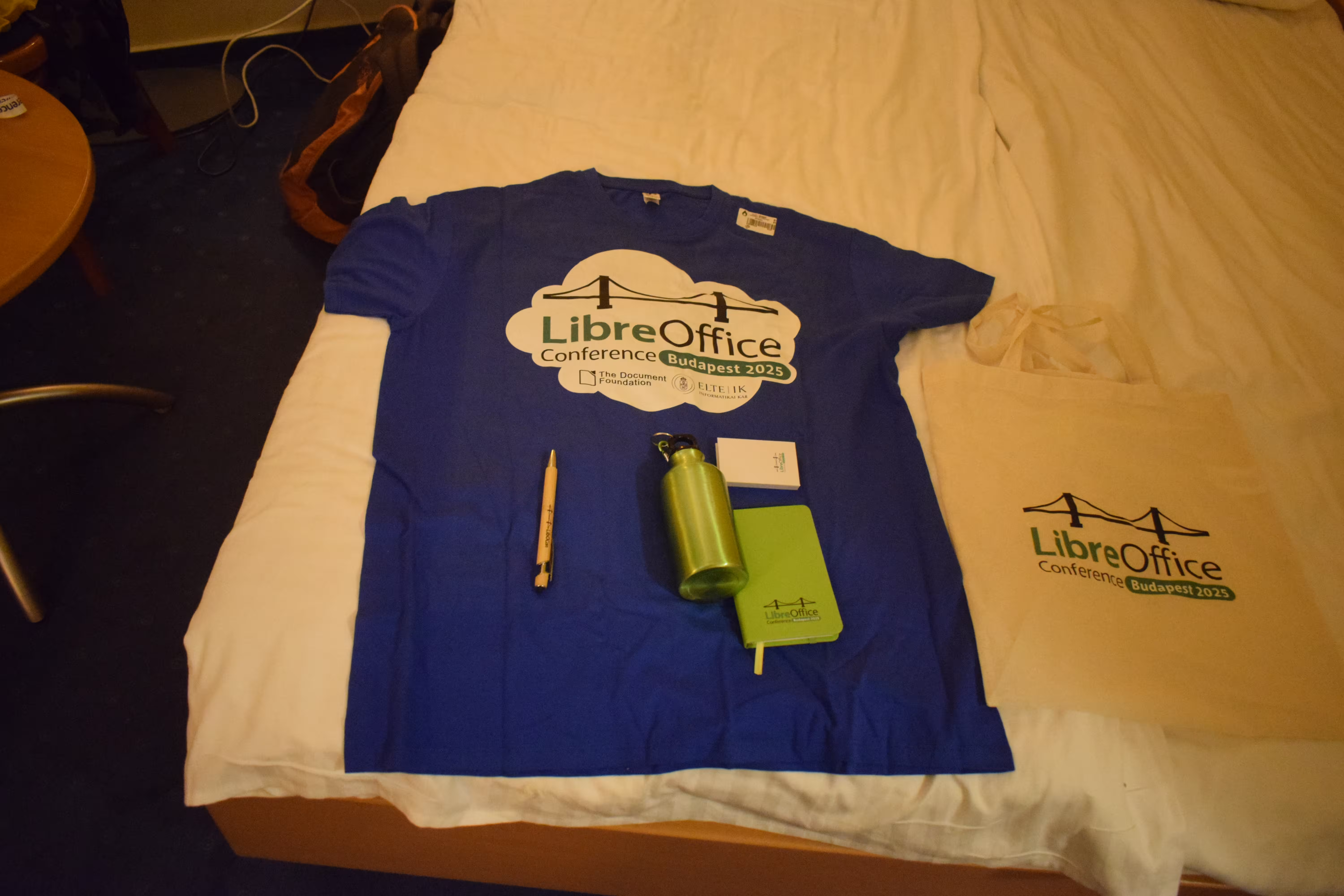 A blue colored T-shirt on a bed along with a pen, a bottle, a diary and a sticky note