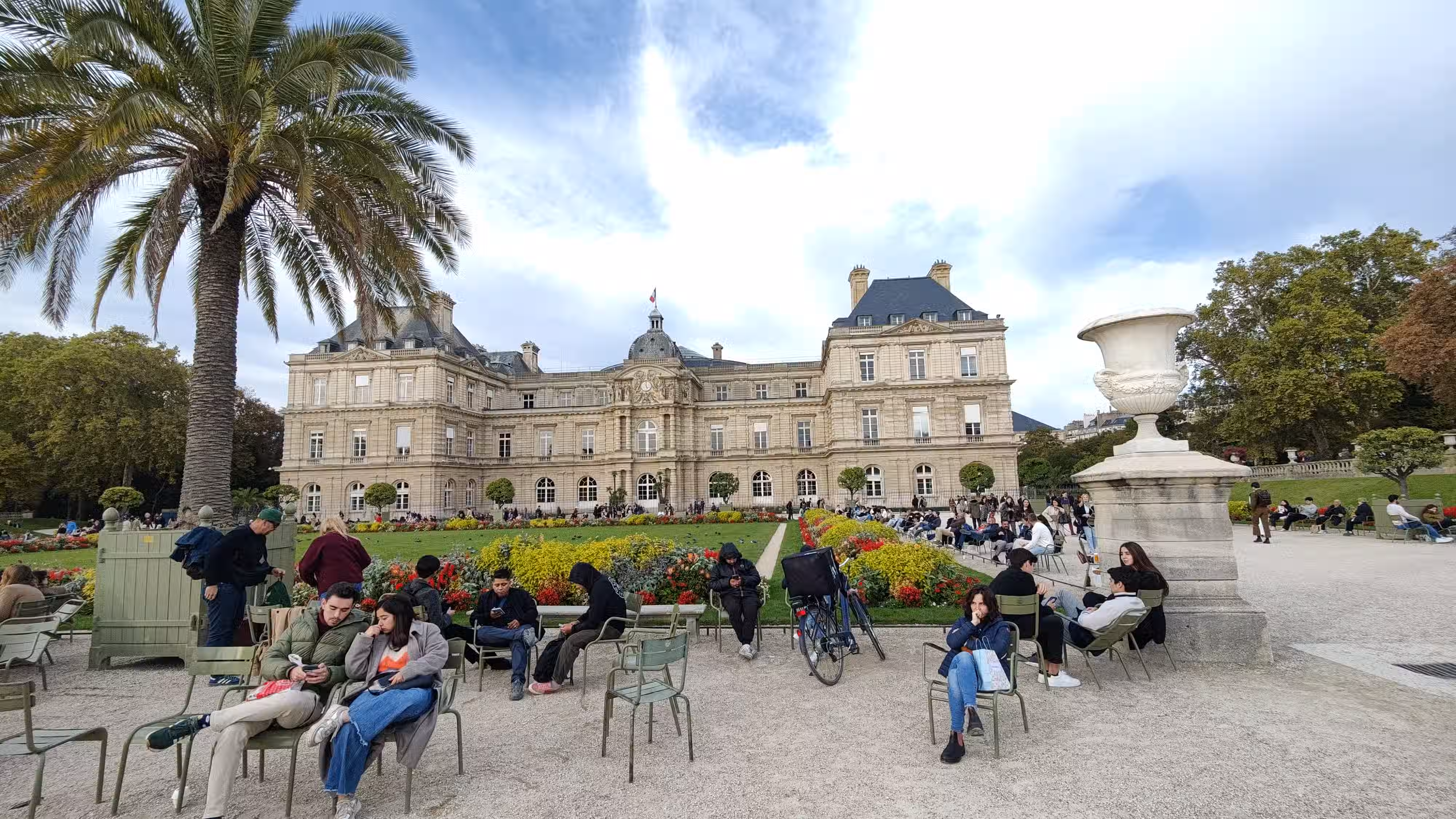 A building with a garden in front it and people sitting closer to us. Sky can be seen in the background.