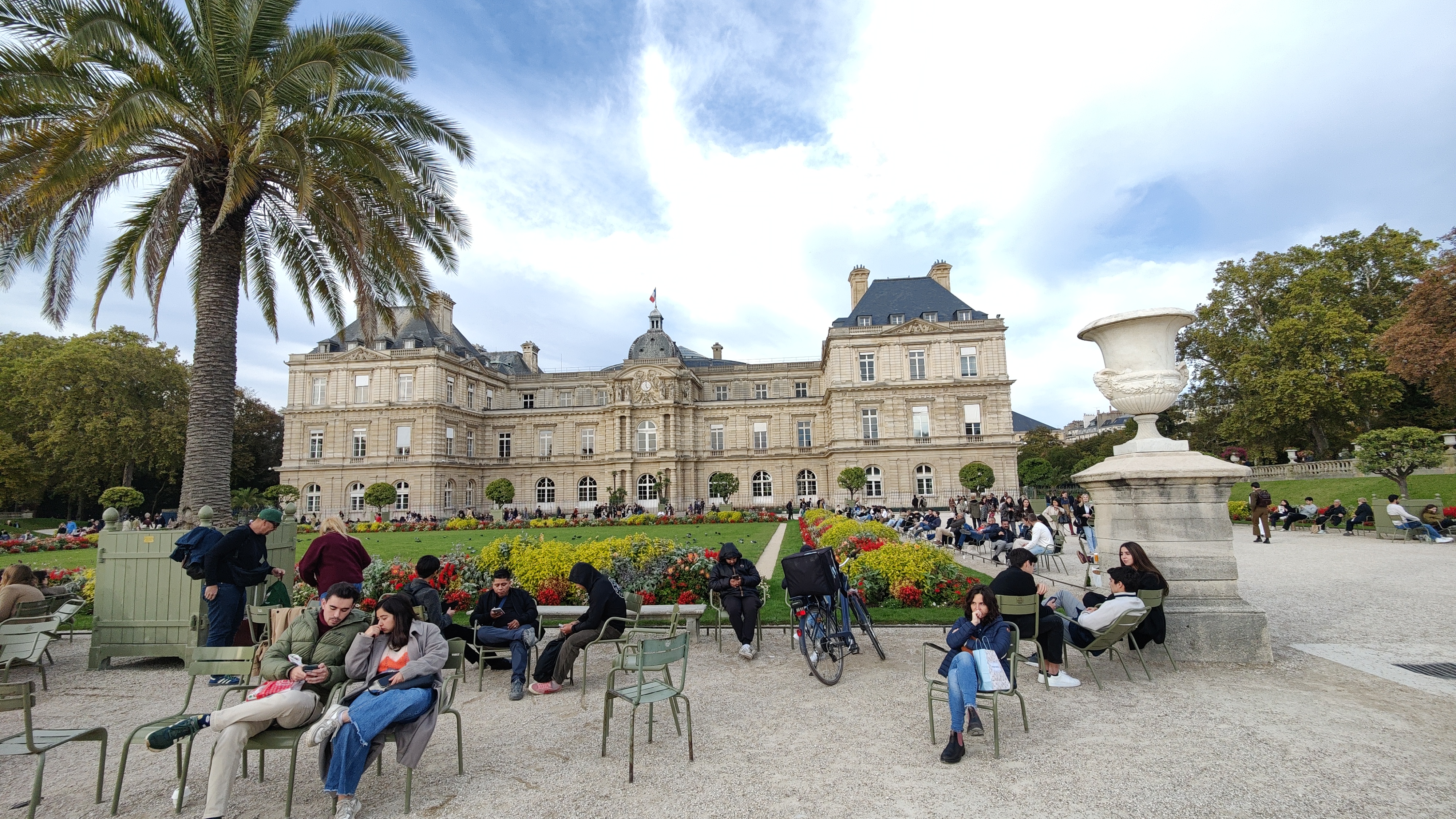 A building with a garden in front it and people sitting closer to us. Sky can be seen in the background.